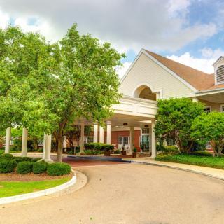 A picturesque building entrance surrounded by lush landscaping and a curved driveway under a partly cloudy sky.