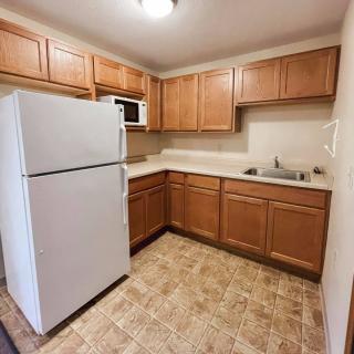 A compact apartment kitchen with wooden cabinets, a refrigerator, microwave, and tiled flooring.
