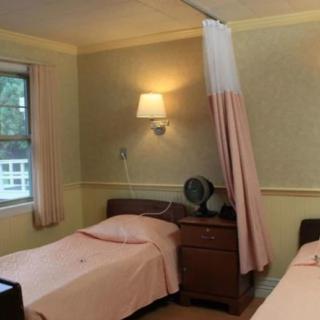 A hospital room with two beds separated by a curtain, featuring a window and a wall-mounted lamp.