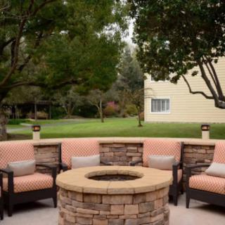 A cozy outdoor seating area with a stone fire pit, surrounded by chairs and lush greenery.
