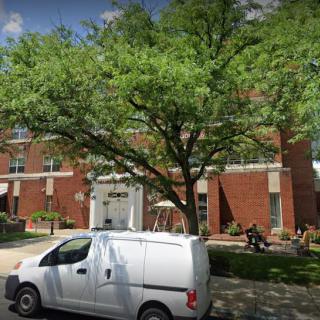 A street view of a brick building with a large tree in front, a white van parked nearby, and people sitting on benches.