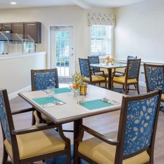 A well-lit dining area with neatly arranged tables and chairs, featuring a view into the kitchen.