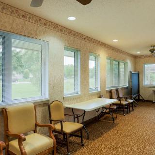 A well-lit waiting room with multiple chairs and tables, featuring large windows and carpet flooring.