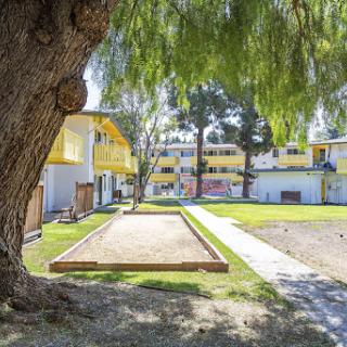 A serene view of an apartment complex featuring yellow buildings, a bocce ball court, and lush green landscaping.