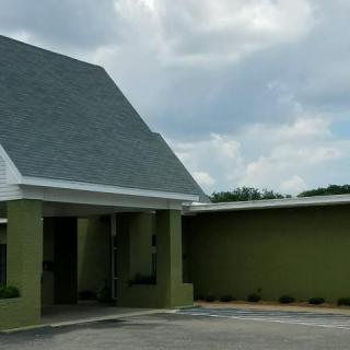 A green building with a sloped roof and a parking area under a cloudy sky.