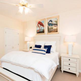 A bright bedroom with white furnishings, featuring a cozy bed, nightstands with lamps, and colorful artwork on the wall.