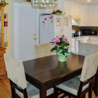 A cozy dining area with a dark wooden table and upholstered chairs, adorned with a floral centerpiece, adjacent to a bright kitchen.