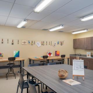 A well-lit art classroom with tables, chairs, and paintings displayed on the wall.