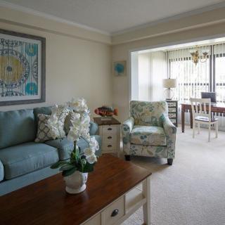 A cozy living room with a blue sofa, patterned armchair, and wooden coffee table. The room is decorated with artwork, cushions, and includes a view of the adjacent dining area.