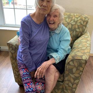 Two elderly women share a joyful embrace while seated indoors.