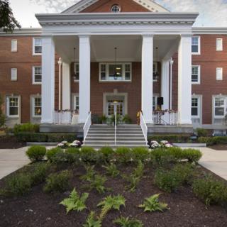 A brick building with tall white columns, landscaped entrance, and steps leading up to the door.