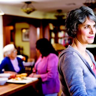 A woman standing in a home setting with others conversing in the background.