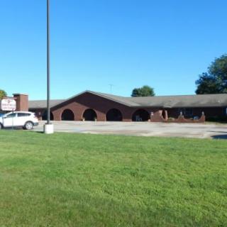 A brick building with arched windows, a parking lot, and a sign in front, under a clear blue sky.