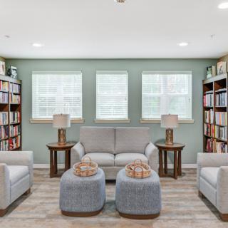 A cozy living room with bookshelves, a sofa, armchairs, and ottomans under a set of three windows.