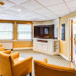 A cozy waiting room with comfortable armchairs and a television, illuminated by natural light through large windows.