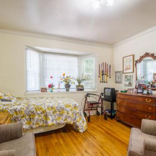 A cozy bedroom with a large window, decorated with plants and vintage furniture on a hardwood floor.