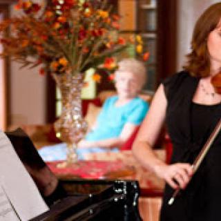A violinist performs in a cozy sitting room, while an elderly listener enjoys the music.