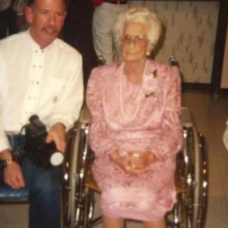 An elderly woman in a pink dress sits in a wheelchair next to a man indoors.