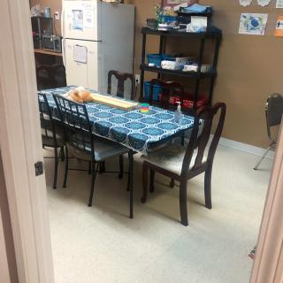 A casual break room with a dining table and chairs, a refrigerator, and shelves.