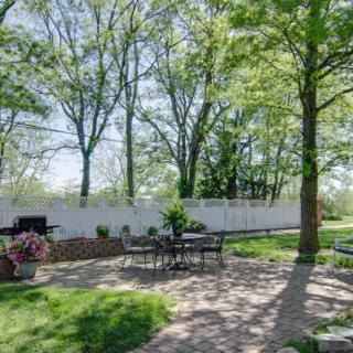 A sunny backyard patio with outdoor seating surrounded by trees and a white fence.