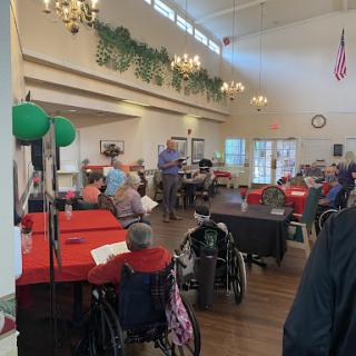 A lively gathering at a community center for seniors, featuring attendees in wheelchairs and festive decorations.