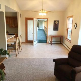 A cozy living room with carpeted floors, featuring a table with chairs and large windows allowing natural light.