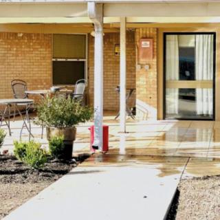 A building entrance featuring a patio area with chairs and tables, surrounded by plants and a walkway.