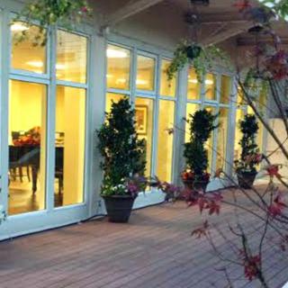 A warmly lit patio with large windows and potted plants on a wooden deck.