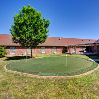 A view of a well-manicured putting green surrounded by a tree and residential building under a clear blue sky.