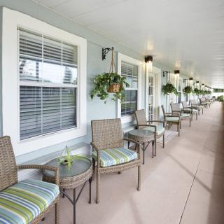 A long patio with multiple wicker chairs and small tables, adorned with hanging plants and windows along the wall.