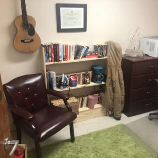 A cozy office space featuring a bookshelf filled with books, a comfortable chair, and a guitar on the wall.