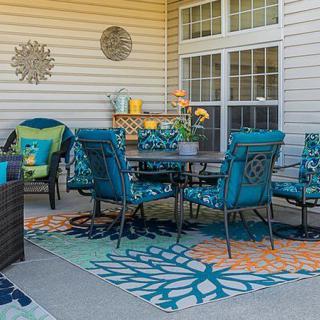 A cozy patio setup with teal cushioned chairs, a table, patterned rugs, and potted plants.
