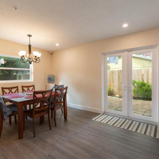 A bright dining room with a wooden table and chairs, featuring a chandelier and glass doors leading to the garden.