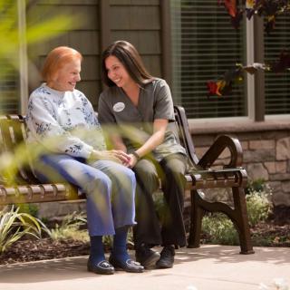 A caregiver and an elderly person share a moment on a bench outside.