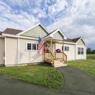 A single-story house with an American flag on the porch, set against a cloudy sky with a neatly maintained lawn.