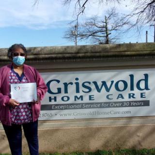 A person wearing a mask holds a certificate in front of the Griswold Home Care sign.