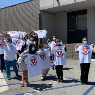 A group of people wearing masks, holding and displaying t-shirts with a logo, standing outside a building.