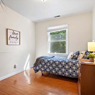 A cozy bedroom with a single bed, adorned with patterned bedding and decorative pillows, features a window allowing natural light to illuminate the hardwood floor.