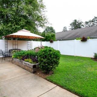 A cozy backyard featuring a patio with outdoor seating and an umbrella, surrounded by lush greenery and a white fence.