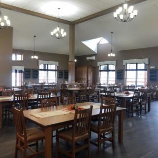 An empty dining hall with wooden tables and chairs, featuring large windows and overhead lighting.