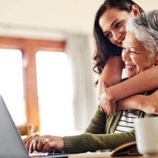 A young woman hugs an elderly woman as they both look at a laptop, sharing a moment of technology and connection at home.