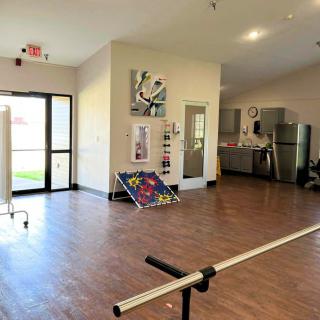 An interior view of a therapy room with rehabilitation equipment, a kitchenette, and wooden flooring.