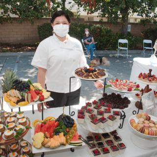 A chef stands by a beautifully arranged buffet table filled with desserts and fruits at an outdoor event.