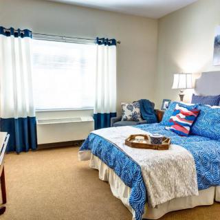 A cozy bedroom with a blue and white color scheme, featuring a neatly made bed, decorative pillows, a bedside lamp, and curtains framing the window.