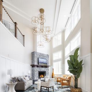 A spacious living room with high ceilings, featuring a modern chandelier, stone fireplace, and large windows that allow for ample natural light.