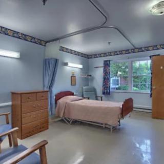 A clean and organized hospital room with a single bed, a dresser, chairs, and a window providing natural light.