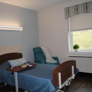 A well-lit hospital room featuring a patient bed, an armchair, and a window with natural light.