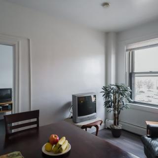 A cozy dining area with a table, fruit bowl, and a television, warmly lit by natural light from a large window.