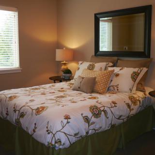 A cozy bedroom with floral bedding, a lamp on the nightstand, and a large mirror above the bed.