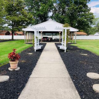 A charming gazebo at the end of a paved walkway surrounded by lush green grass and trees.